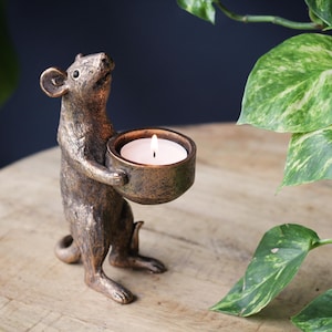 May include: A bronze-colored mouse figurine standing on its hind legs and holding a small, round bowl. The bowl contains a lit white tea light candle. The mouse has a textured, metallic finish with dark highlights and a pointed snout.
