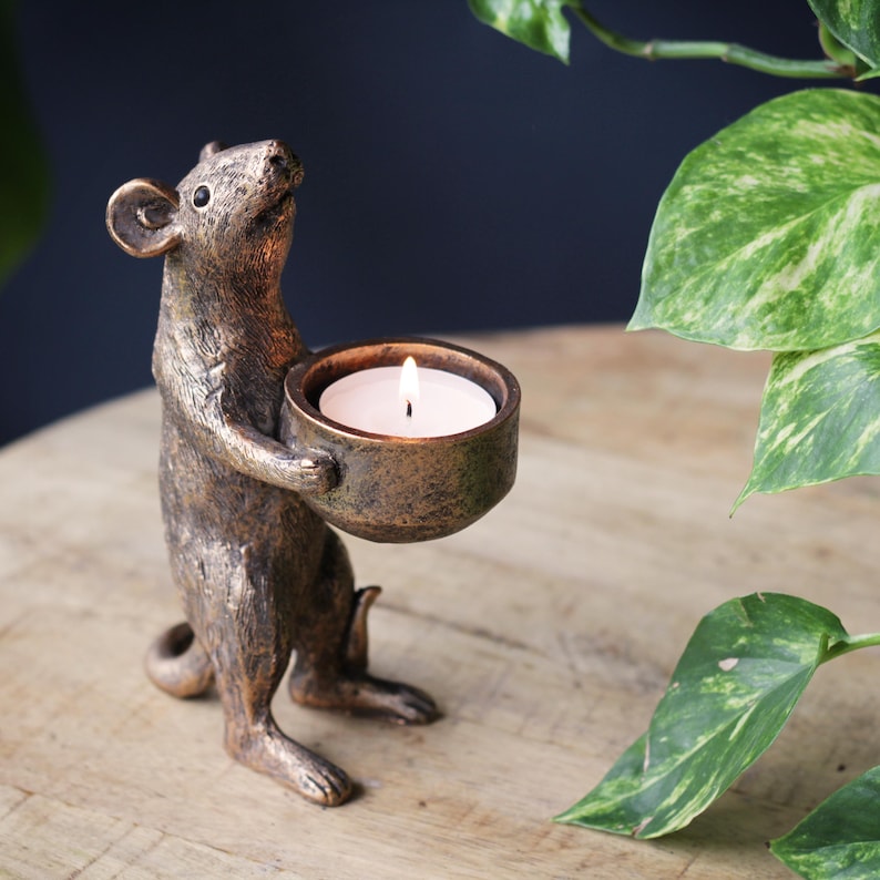 May include: A bronze-colored mouse figurine standing on its hind legs and holding a small, round bowl. The bowl contains a lit white tea light candle. The mouse has a textured, metallic finish with dark highlights and a pointed snout.