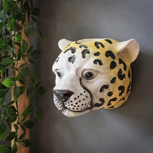 Puede incluir: Escultura mural detallada de la cabeza de un jaguar. La escultura presenta una cara color crema con manchas negras y una corona amarillo-marrón. Los ojos del jaguar son oscuros y realistas, y la nariz es negra. La escultura está montada en una pared gris.