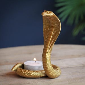 May include: A gold cobra-shaped candle holder with a lit white tea light candle. The cobra is in an upright pose, with detailed scales and a textured surface. The candle holder is on a wooden surface.