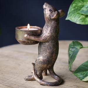 May include: A bronze-colored mouse figurine standing on its hind legs, holding a small, round bowl in its front paws. The bowl contains a lit white tea light candle. The mouse has a textured, metallic finish with dark highlights and a long, curved tail.