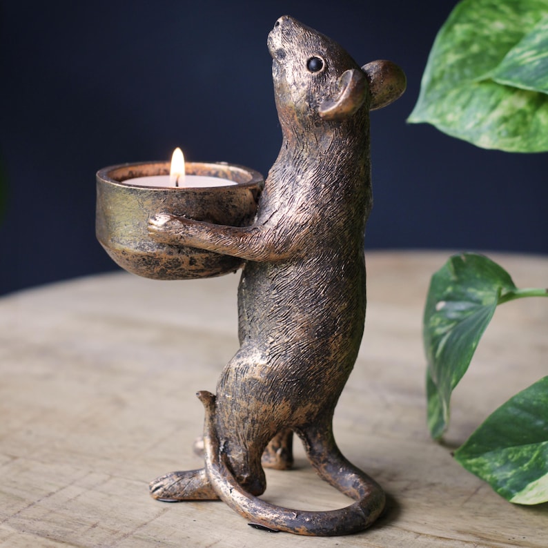 May include: A bronze-colored mouse figurine standing on its hind legs, holding a small, round bowl in its front paws. The bowl contains a lit white tea light candle. The mouse has a textured, metallic finish with dark highlights and a long, curved tail.