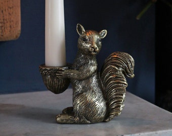 Pode incluir: Castiçal de esquilo dourado com uma vela branca. O esquilo está segurando uma bolota em suas patas.