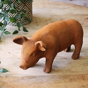 Op de afbeelding: Een terracotta-kleurige varkensbeeldje met een textuur. Het varken staat met zijn hoofd naar beneden. De oren staan omhoog en de snuit is iets puntig. Het beeldje is een decoratief item.