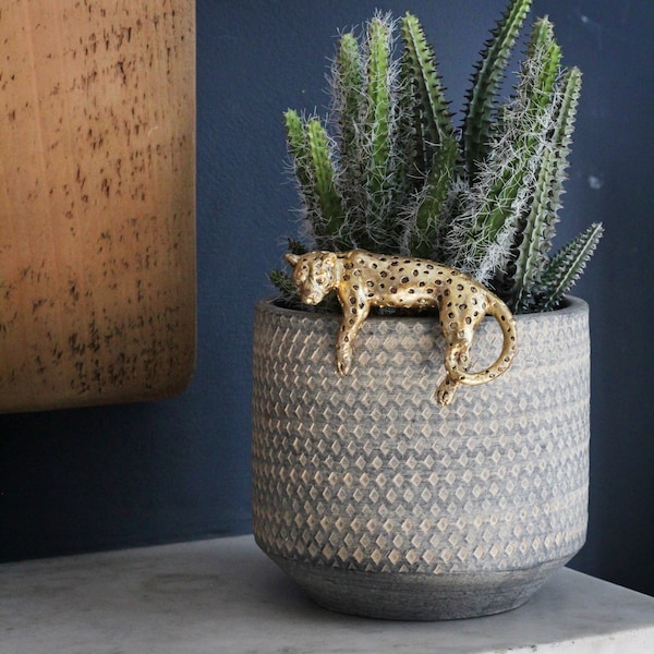 May include: A gold-colored leopard figurine perched on the edge of a gray planter with a diamond pattern. The planter is filled with a green cactus plant.