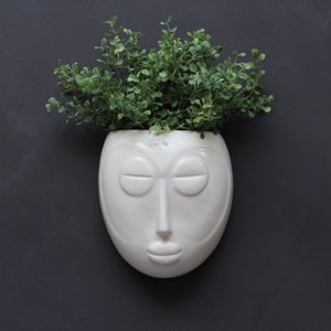 May include: White ceramic planter shaped like a face with closed eyes and a closed mouth, with a green plant growing out of the top.
