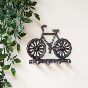 Op de afbeelding: Een donkergrijze metalen wandhaak in de vorm van een fiets. Het fietsframe heeft twee wielen met spaken en een zadel. Onder de fiets bevindt zich een horizontale balk met vier afgeronde haken voor het ophangen van voorwerpen.