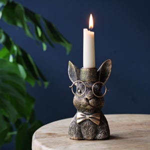 May include: A bronze-colored rabbit-shaped candle holder with a lit white candle. The rabbit wears round glasses and a bow tie. The candle holder is on a wooden surface with a dark blue background.