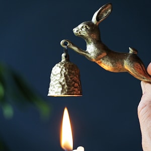 May include: A brass rabbit ornament holding a bell, with a lit candle beneath. The rabbit is depicted mid-jump, with the bell hanging from its paw. The bell has a textured surface. The background is a dark blue.