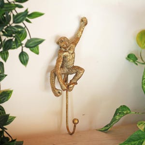May include: A gold-colored monkey-shaped wall hook. The monkey is depicted in a climbing pose, with one arm raised. The hook is attached to a long, slender rod ending in a rounded hook. The background is a neutral wall with green foliage.