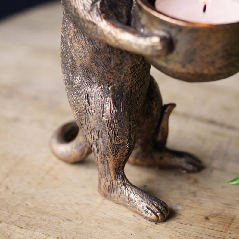 May include: A bronze-colored animal figurine standing on its hind legs, featuring a textured fur-like finish. The figure holds a small, round bowl in its front paws, which contains a white tea light candle with a visible lit wick.