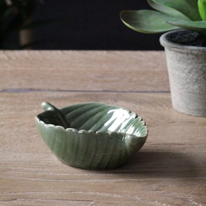 Puede incluir: Un plato de cerámica verde en forma de hoja con una superficie texturizada. El plato es perfecto para guardar objetos pequeños como joyas o llaves.