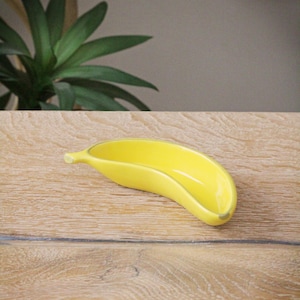 Puede incluir: Un plato de cerámica amarillo con forma de plátano. El plato está sobre una superficie de madera marrón claro.