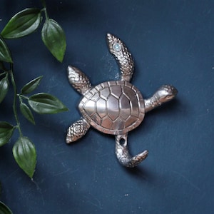 May include: A silver-toned metal wall hook shaped like a sea turtle. The turtle features a textured shell, detailed flippers, and a small embedded blue crystal on one flipper. The bottom flipper is curved upward to function as a hook for hanging items.