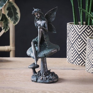 Op de afbeelding: Een bronzen fee-figuur dat op een paddenstoel zit met een bloemenkrans en vleugels. De fee houdt een bloem in haar handen. De paddenstoel heeft een bruine steel en een groene dop.