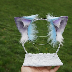 Puede incluir: Un par de orejas de gato azules y moradas con ribete de piel blanca, unidas a una diadema negra. Las orejas están en un expositor blanco.