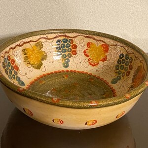 May include: A large, round ceramic bowl with a cream-colored base and a green, brown, and red rim. The bowl is decorated with a repeating pattern of grapes and leaves in shades of green, blue, red, and yellow.