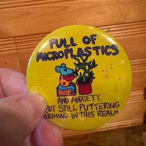 May include: A yellow button with the text "FULL OF MICROPLASTICS AND ANXIETY. BUT STILL PUTTERING AROUND IN THIS REALM." A cartoon mouse is tending a plant. The button is held by a person.