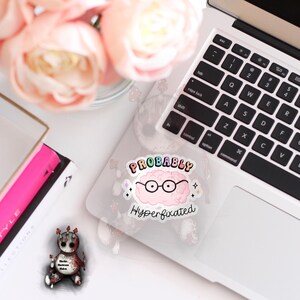 May include: A sticker featuring a pink brain wearing glasses and the text "PROBABLY Hyperfixated" in rainbow lettering.