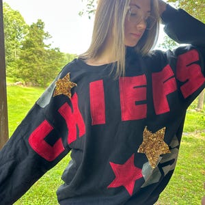May include: Black sweatshirt with the word "CHIEFS" in red lettering. The sweatshirt has two star-shaped patches, one red and one gold, and a camouflage accent on the sleeve.