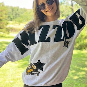 May include: A light gray sweatshirt with the word "MIZZOU" in large black letters across the chest. The sweatshirt has a star patch and a patch that says "MIZZOU Missouri". The person is wearing black shorts and sunglasses.