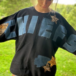 May include: Black sweatshirt with the word "CHIEFS" appliquéd in denim and camouflage fabric. Gold and camouflage star accents are also sewn onto the sweatshirt. The sweatshirt is a casual, oversized style.