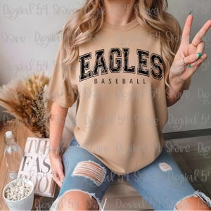 Puede incluir: Una camiseta color beige con el texto "EAGLES BASEBALL" en negro y con un efecto desgastado. La camiseta se combina con unos vaqueros azules rotos. La imagen tiene una estética casual y deportiva.
