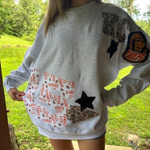 May include: A gray sweatshirt with patches of leopard print, black stars, and a Kansas City Chiefs logo. The sweatshirt has a patch with the word "Kansas City" and footballs.
