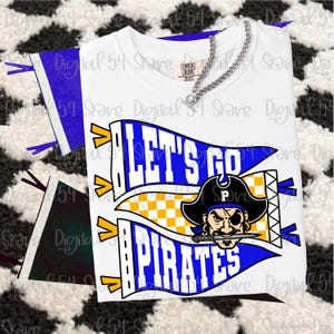 Pode incluir: Camiseta branca com um design gráfico com as palavras "LET'S GO PIRATES" em azul e branco em uma flâmula. O design inclui um pirata com chapéu preto e espada. Um colar de corrente de prata está sobre a camiseta.