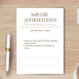 May include: A white card with the text "Soft Life Affirmations" in a serif font. The card includes affirmations about self-worth and taking up space. A gold and white pen rests on the left side of the card.