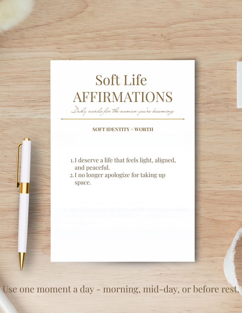 May include: A white card with the text "Soft Life Affirmations" in a serif font. The card includes affirmations about self-worth and taking up space. A gold and white pen rests on the left side of the card.