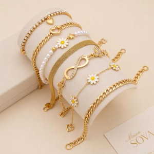 May include: A selection of gold-coloured bracelets with diverse designs, including hearts, stars, daisies, an infinity symbol, and chain links. Some bracelets feature clear gemstones and white pearls. The bracelets are displayed on a white surface.