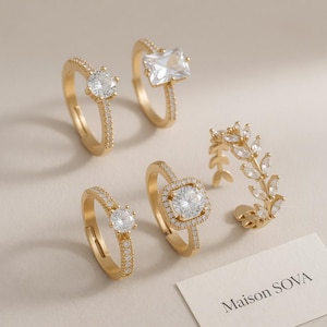 Peut inclure: Plusieurs bagues dorées ornées de pierres précieuses claires. Les modèles comprennent une pierre carrée, un sertissage halo et une bague en forme de feuille. Une carte avec le texte "Maison SOVA" est visible.