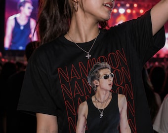 Op de afbeelding: Een zwart T-shirt met korte mouwen en een herhalend rood tekstpatroon met de tekst NAMJOON. Het midden van het shirt toont een afbeelding van een persoon in een zwart hemd, een zonnebril en gelaagde zilveren kettingen over de rode tekst.