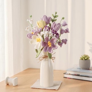 May include: A handmade bouquet of crocheted flowers in a white vase. The bouquet features purple, yellow, and white flowers with green stems and leaves. The vase is tied with a twine bow and sits on a small, square, woven mat.