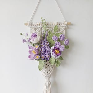 May include: A cream-coloured macrame wall hanging with a crocheted floral arrangement. The flowers are in shades of purple, lavender, and white, with green leaves. The macrame has a wooden dowel and hanging cord, with a tassel at the bottom.