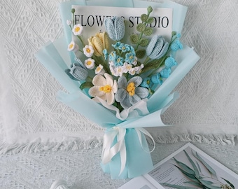May include: A crocheted bouquet of blue and yellow flowers wrapped in light blue paper with a white ribbon. The bouquet is sitting on a white textured surface with a magazine titled "Floral Fancy" in the foreground.