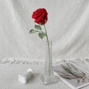 May include: A handmade red crochet rose with green leaves in a clear glass vase. The vase is shaped like the Eiffel Tower. A small white decorative column and a magazine are also in the image.