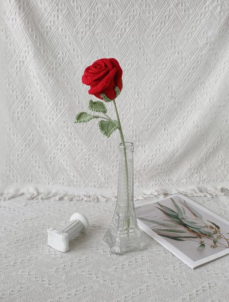 May include: A handmade red crochet rose with green leaves in a clear glass vase. The vase is shaped like the Eiffel Tower. A small white decorative column and a magazine are also in the image.