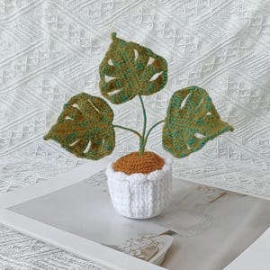 Op de afbeelding: Een handgemaakte gehaakte plant met drie groene en bruine bladeren. De plant staat in een witte gehaakte pot met een bruine bovenkant. De plant wordt gepresenteerd op een wit oppervlak.
