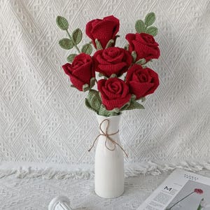 May include: A bouquet of six red crocheted roses with green leaves in a white vase tied with twine. The vase sits on a white surface with a decorative column and a magazine.