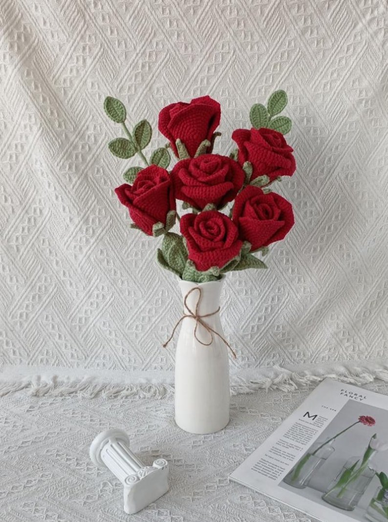 May include: A bouquet of six red crocheted roses with green leaves in a white vase tied with twine. The vase sits on a white surface with a decorative column and a magazine.