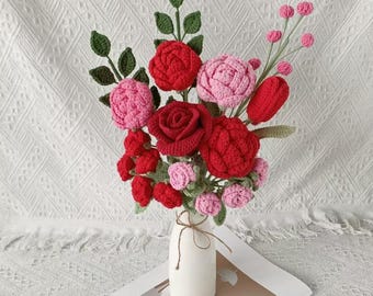May include: A handcrafted bouquet of crochet flowers in a white vase. The bouquet features red and pink roses, buds, and green leaves. The vase is tied with twine and sits on a white surface with a decorative paper.