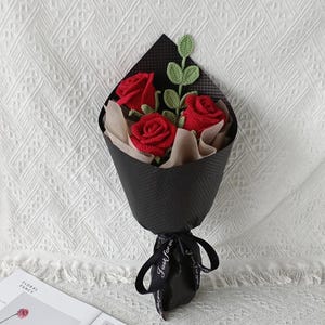 May include: A bouquet of three red crocheted roses with green leaves, wrapped in black paper and tied with a black ribbon. The ribbon has the text "Just for you" printed on it. The bouquet is presented on a white textured surface.