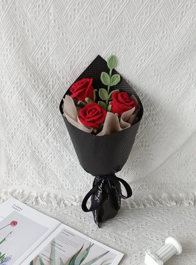 May include: A bouquet of three red crocheted roses with green leaves, wrapped in black paper and tied with a black ribbon. The ribbon has the text "Just for you" printed on it. The bouquet is presented on a white textured surface.