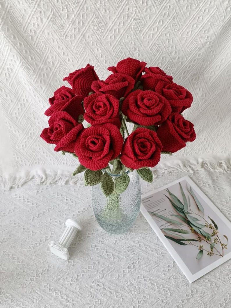 May include: A bouquet of red crocheted roses in a clear glass vase. The handmade flowers have green leaves and stems. A small white decorative column and a magazine with botanical illustrations are also in the image.