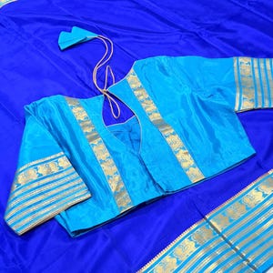 May include: A vibrant royal blue saree featuring a wide border with gold metallic floral patterns and horizontal stripes. The matching light blue blouse has a deep back neckline with thin gold ties and gold floral trim along the edges.