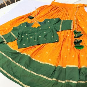 May include: A two-piece outfit consisting of a bright orange pleated skirt and a dark green blouse. The skirt features a wide dark green hem with gold metallic stripes and decorative hanging tassels. The green blouse has a scalloped neckline.