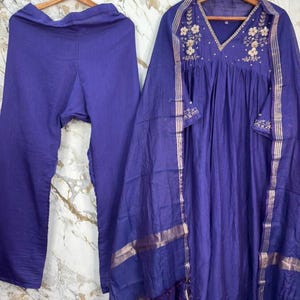 May include: A deep purple three-piece outfit consisting of a pair of trousers, a long-sleeved tunic with a V-neckline featuring floral embroidery, and a matching sheer dupatta with metallic gold-toned striped borders hanging on wooden hangers.