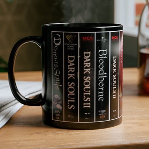 Peut inclure: Mug noir en céramique avec un design inspiré des jeux vidéo. La tasse affiche les titres "Demon's Souls", "Dark Souls", "Dark Souls II" et "Bloodborne" dans un style cassette VHS. De la vapeur s'élève de la tasse.